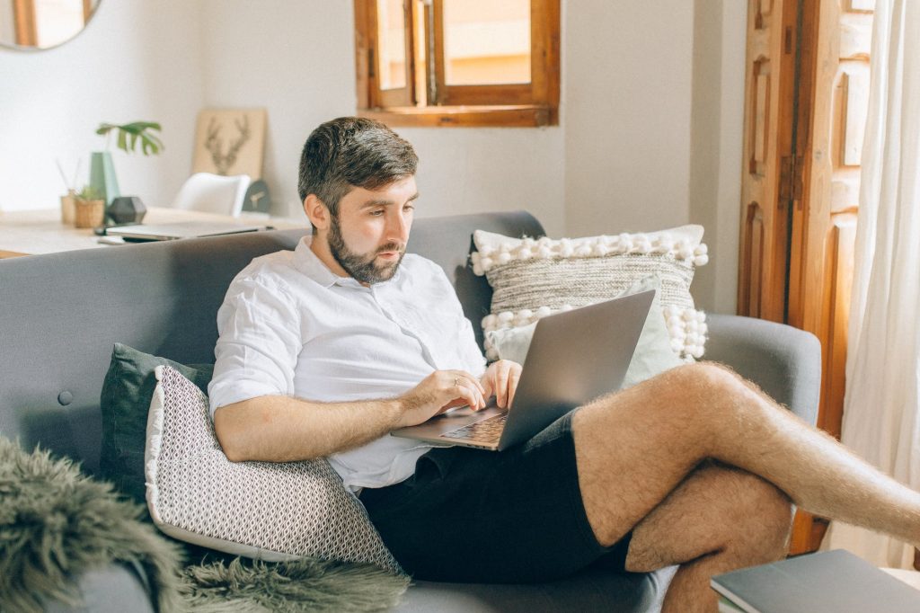 man in white polo shirt and black shorts sitting on gray couch using macbook
